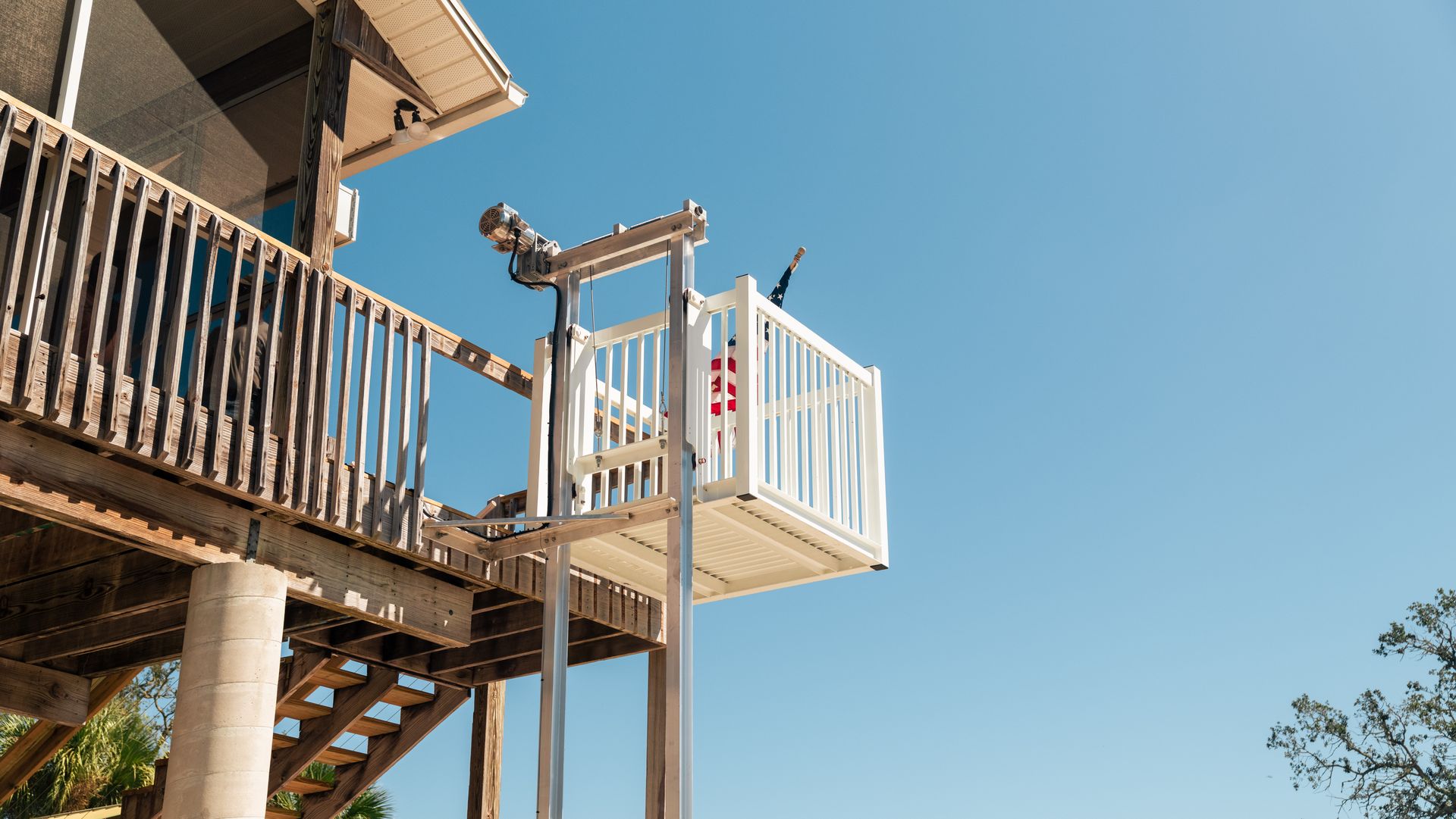 Exterior view of a white platform lift ascending from a wooden deck, person onboard, blue sky.