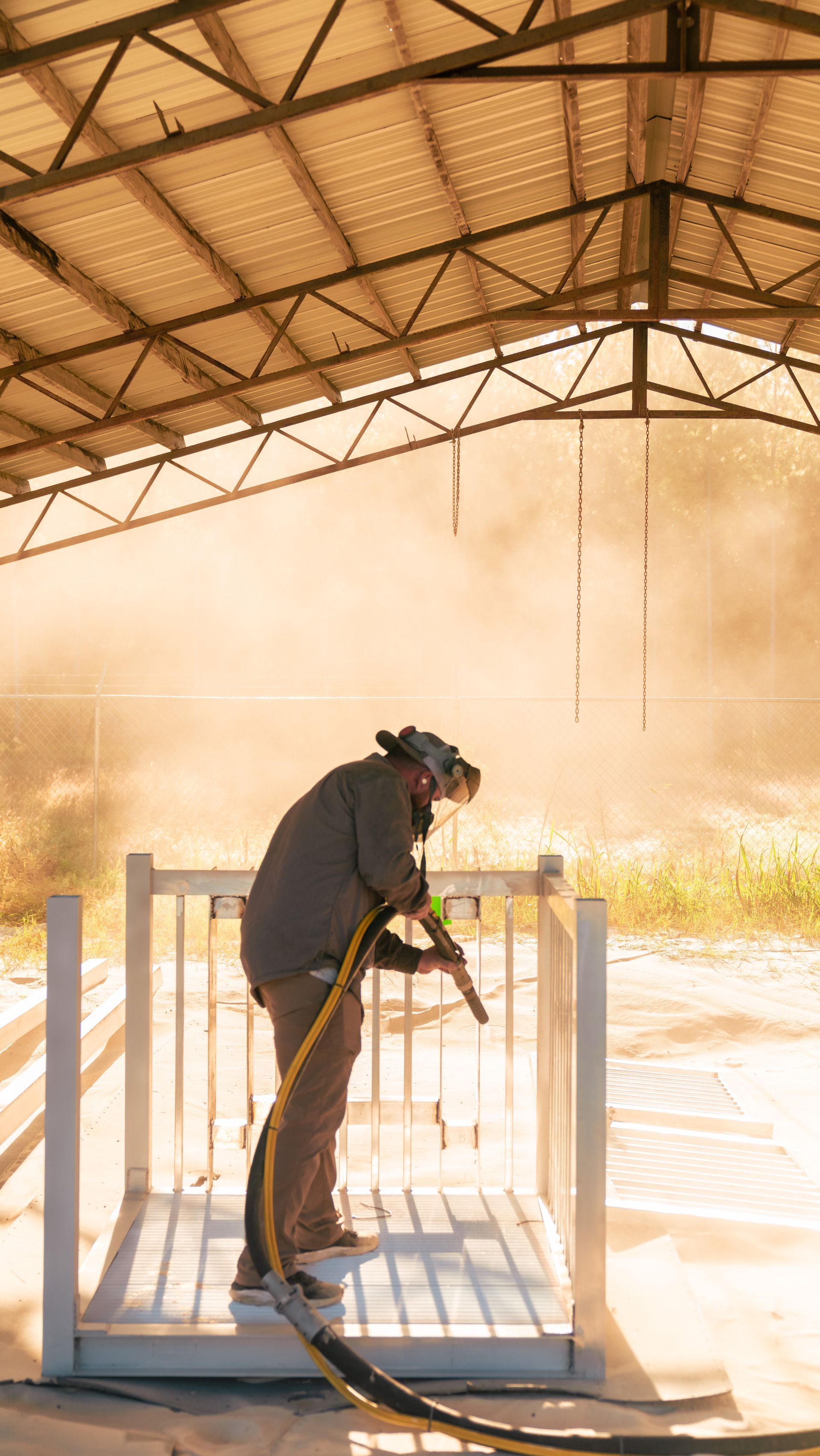 Person sandblasting metal structure in a dusty, open-air workshop, wearing protective gear.