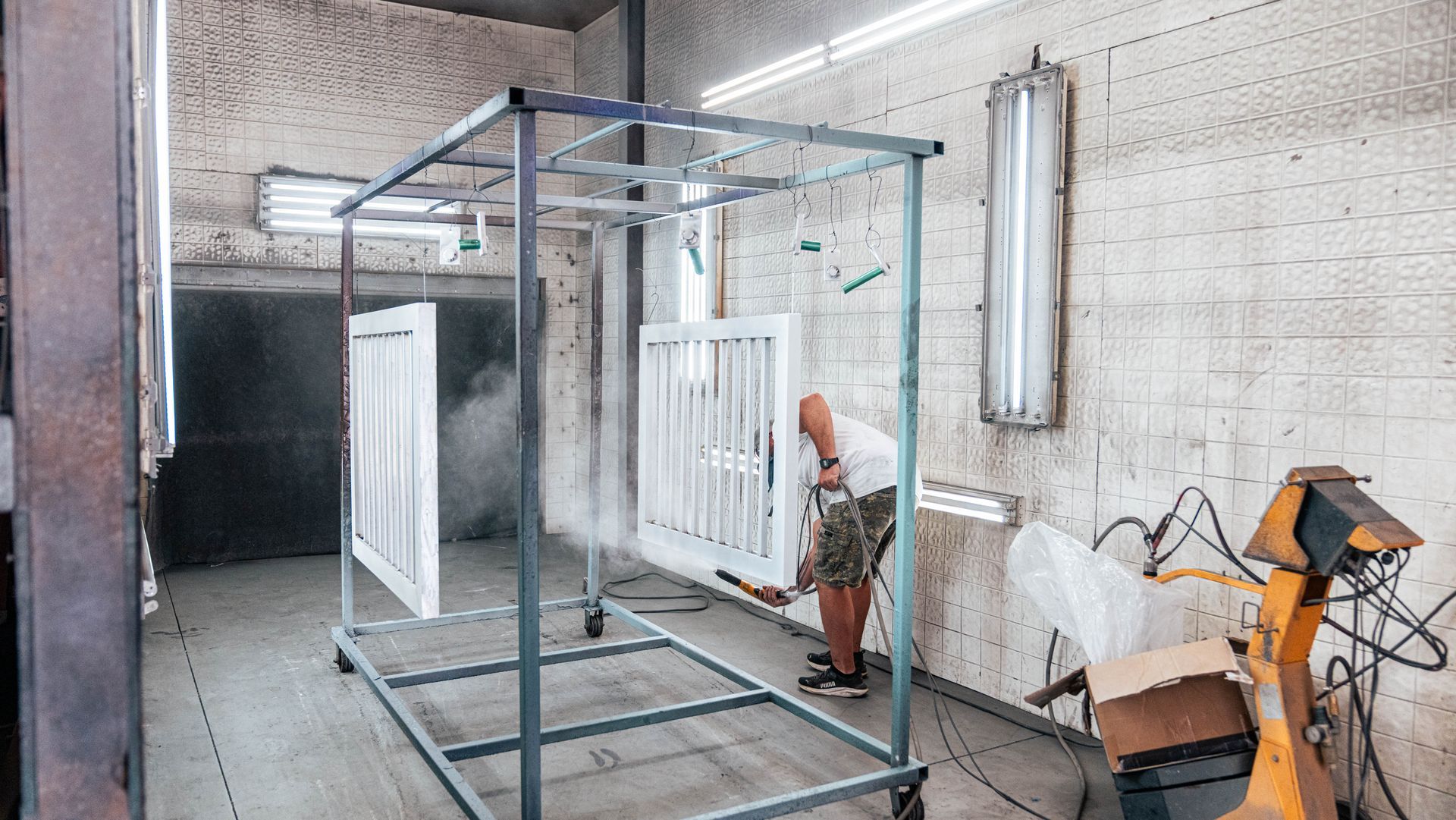 Man spray-painting white gates inside a workshop. Gray metal frame holds gates; white paint fills the air.