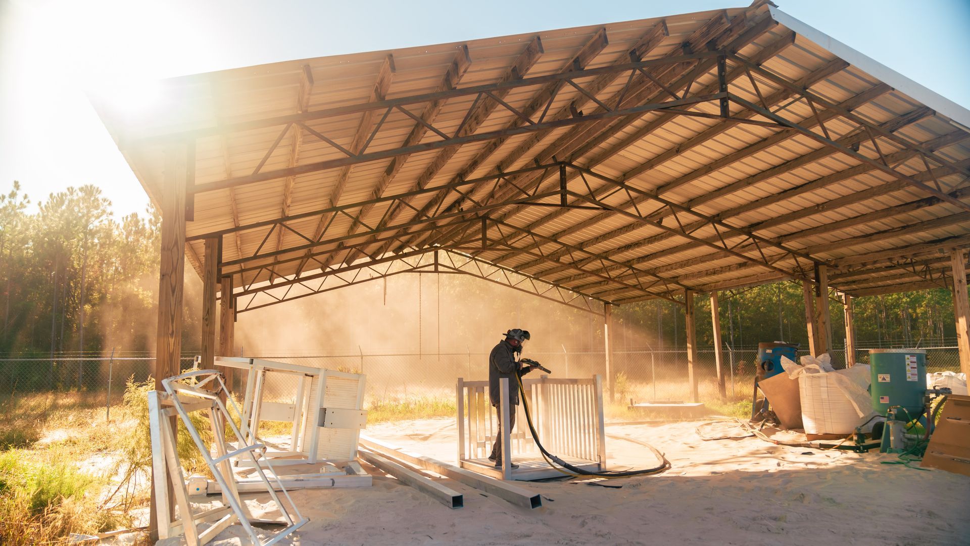 Man sandblasting metal framework inside a large open-sided structure with bright sunlight.