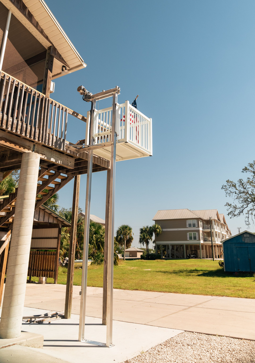 Lift on a stilt house, with a bird perched on top. Buildings and green lawn on a sunny day.