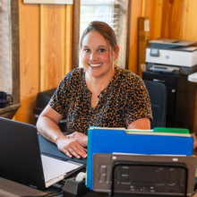 Woman smiling at a desk, wearing a leopard-print top, in a wood-paneled office.