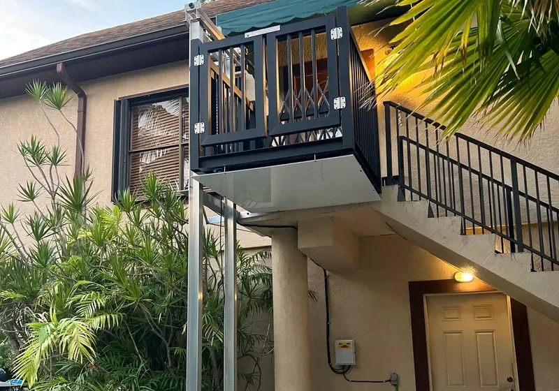 Wheelchair lift installed next to outdoor stairs. Black lift enclosure, beige building, greenery.