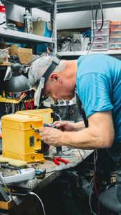 Een persoon in een werkplaats die gereedschap gebruikt op een gele machine, voorovergebogen over een rommelige werkbank.