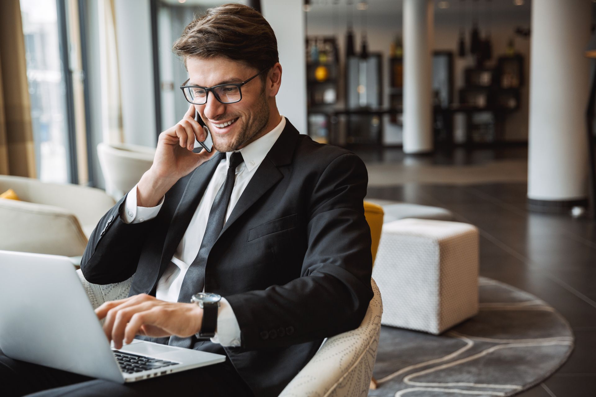 A man in a suit is sitting in a chair using a laptop and talking on a cell phone.