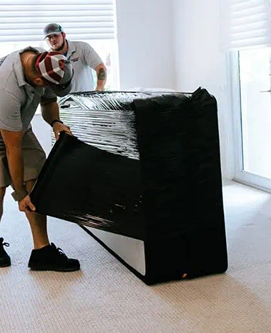 A man is wrapping a black couch in plastic wrap.