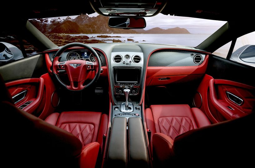 Red leather interior of a Bentley luxury car, with a view of the ocean and mountains through the windshield.
