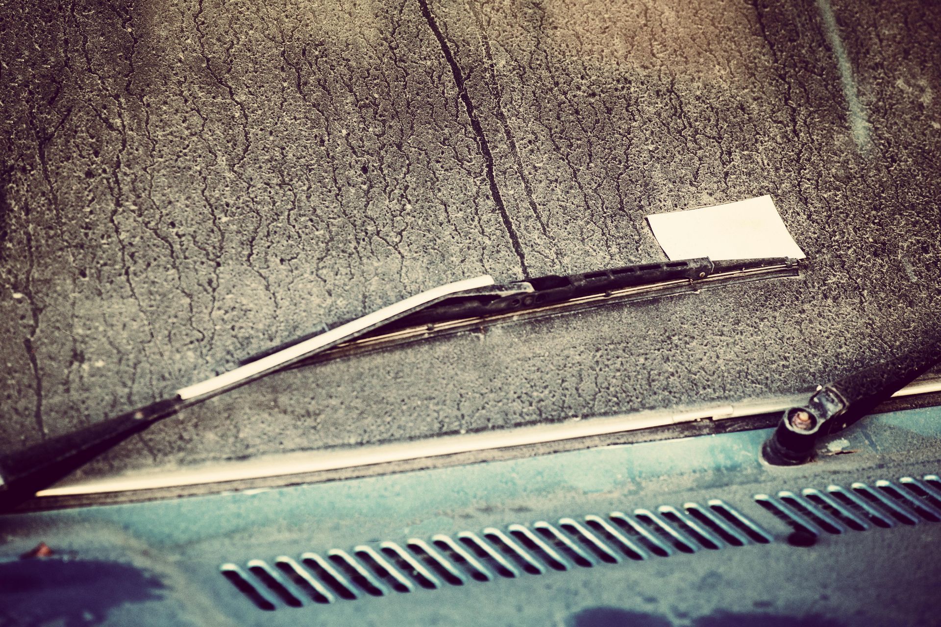 Windshield covered in dirt, a white card tucked under a wiper, on a teal car.