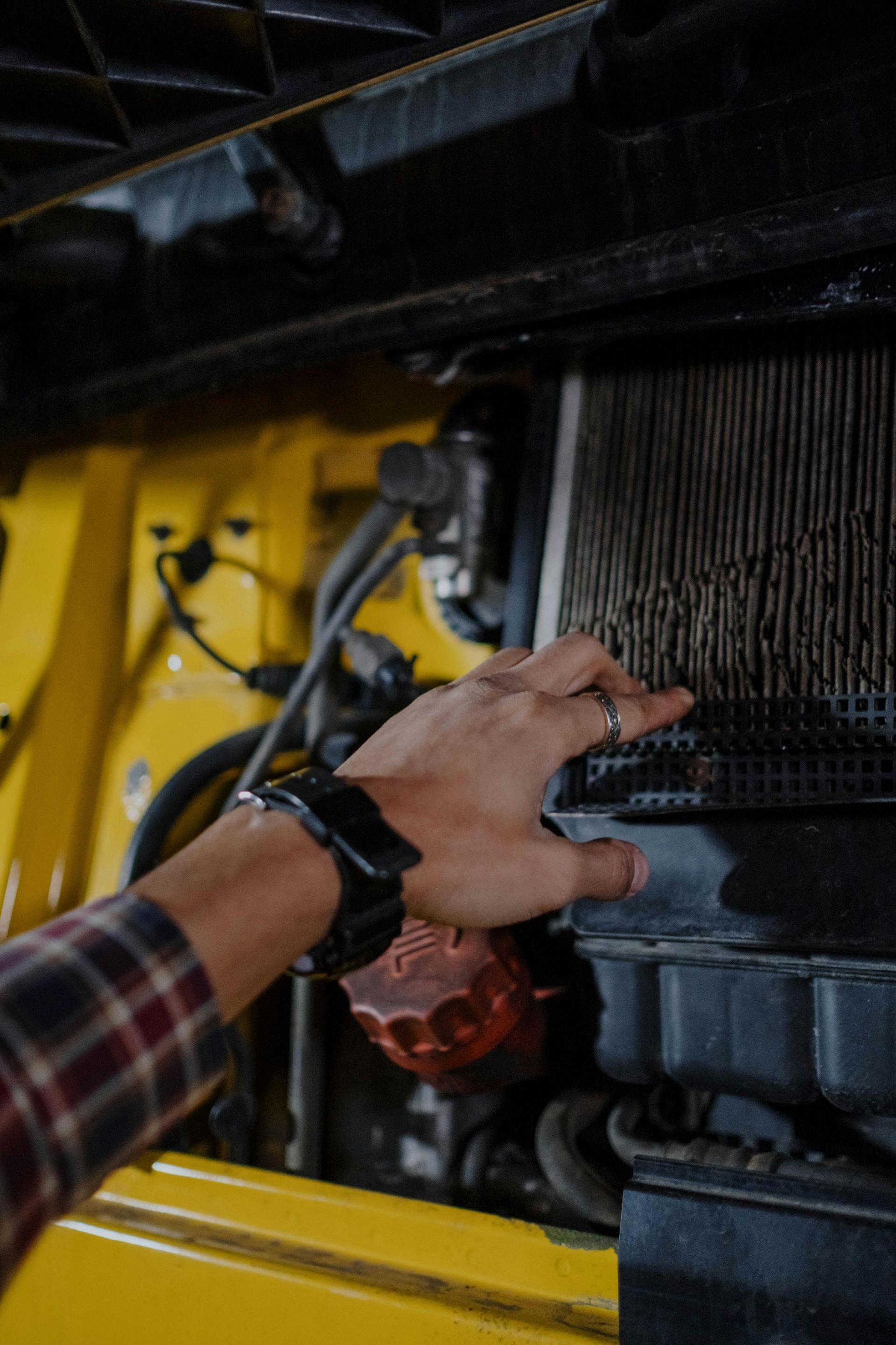 Person's hand pointing to a dirty air filter in a yellow vehicle engine compartment.