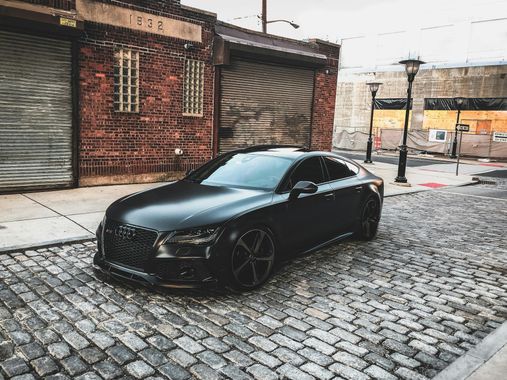 Black car parked on cobblestone street in front of a brick building with closed garage doors.