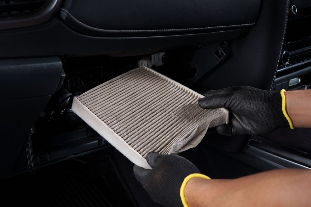 Gloved hands removing a dirty cabin air filter from a car's dashboard.