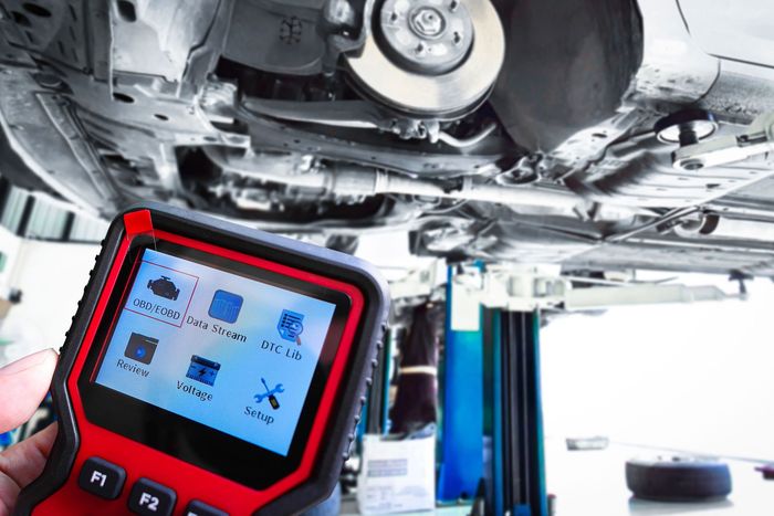 A handheld diagnostic scanner displaying menu icons, held in front of a car raised on a lift in a workshop. A handheld red automotive diagnostic scanner focused on the undercarriage of a vehicle raised on a car lift.