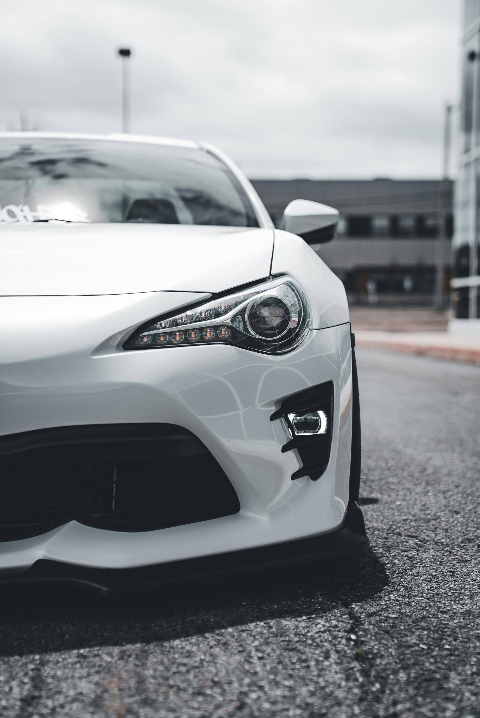 A low-angle, close-up shot of a white sports car’s front end, highlighting its headlight and bumper on an asphalt road.