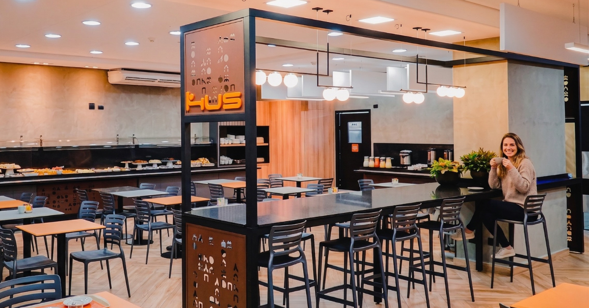 Mulher tomando café da manhã no Hus Hotel Maringá em ambiente confortável e acolhedor.