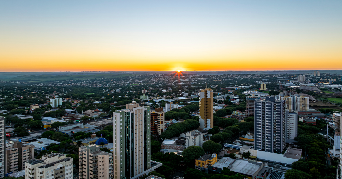 Vista aérea panorâmica de Maringá ao pôr-do-sol, o horizonte da cidade sob uma luz alaranjada.