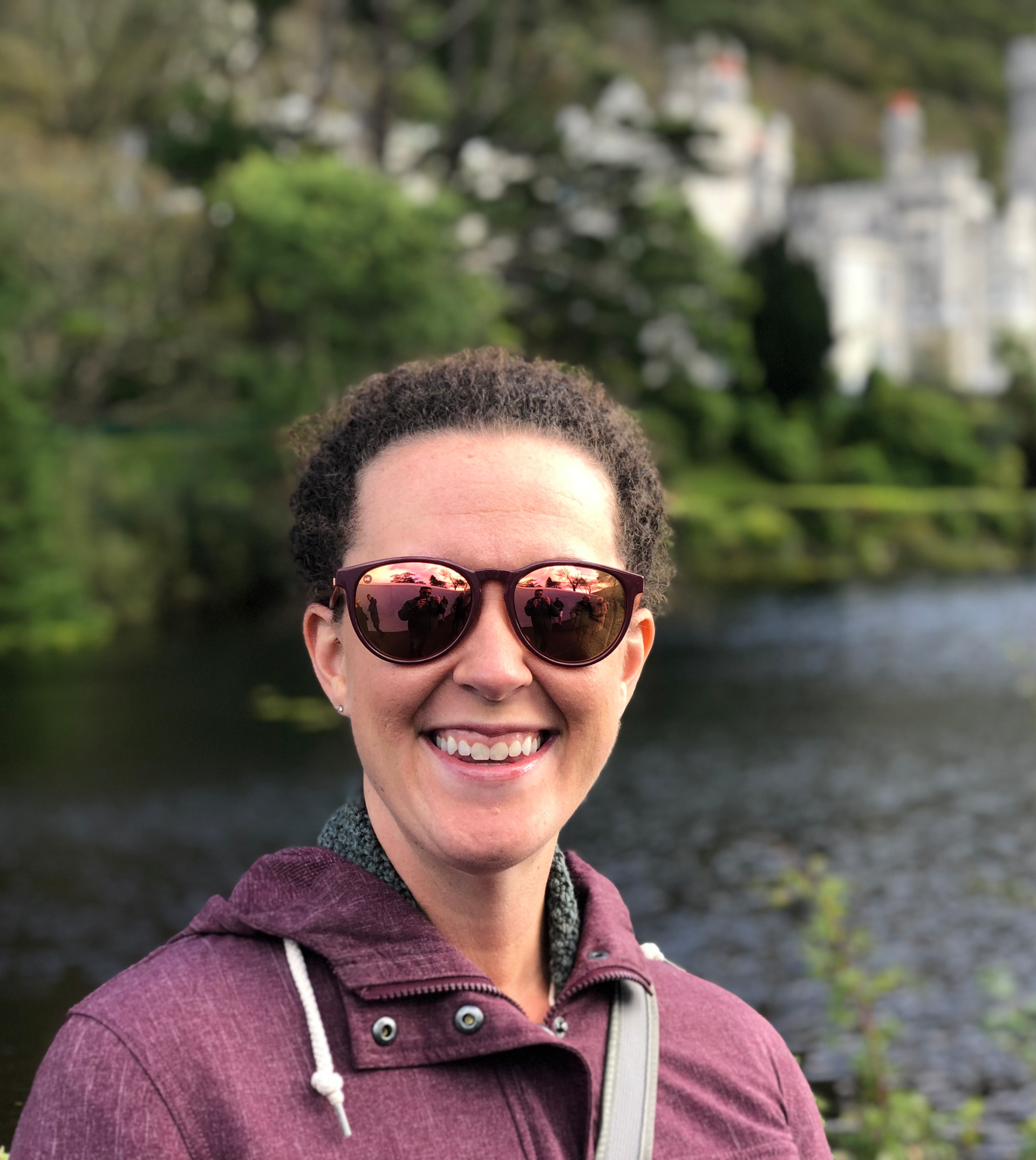 Woman in sunglasses smiles, wearing a burgundy hooded jacket. Background features a lake and blurred white castle.