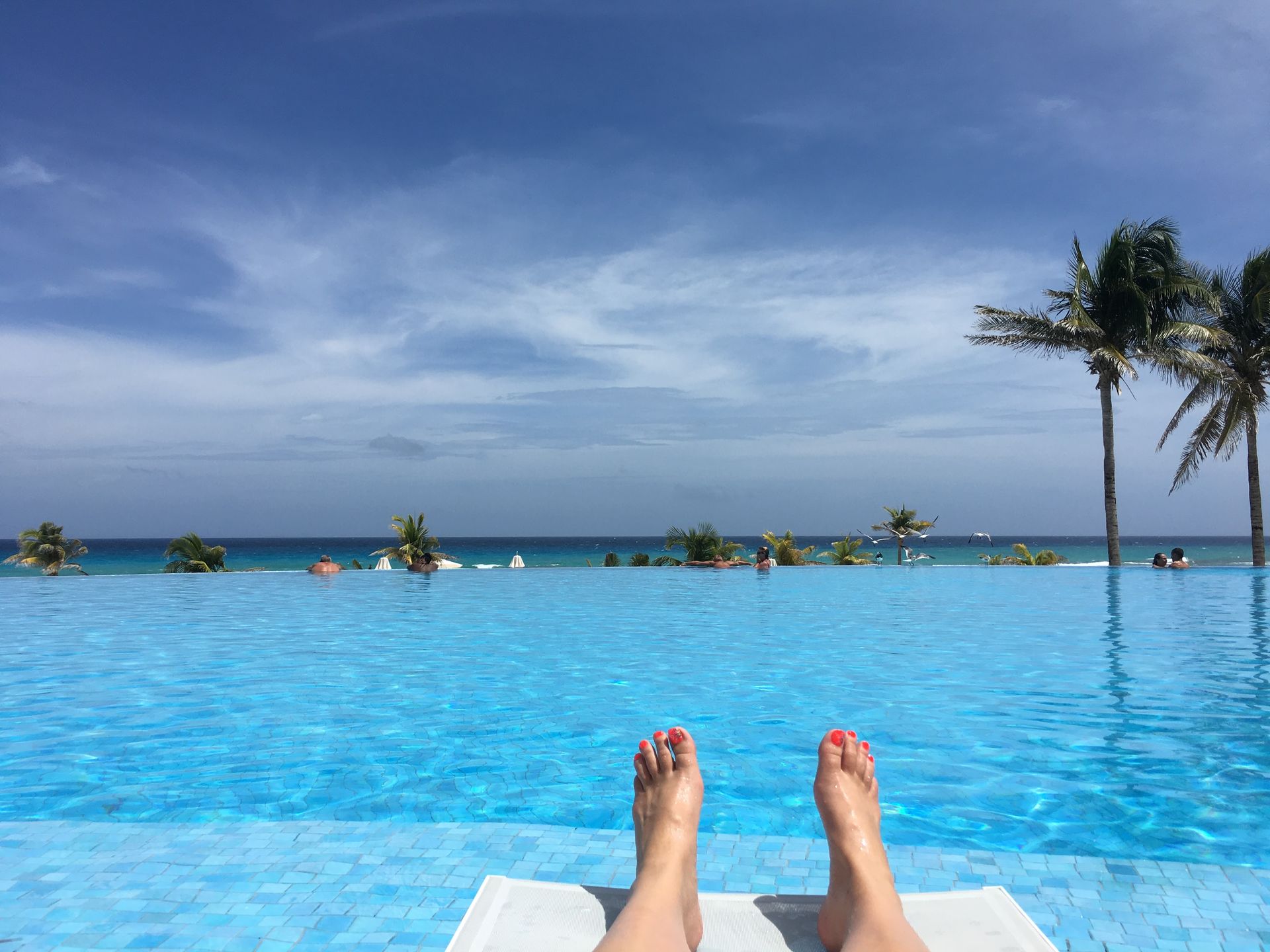 Feet of person sunbathing by a turquoise infinity pool overlooking the ocean, palm trees under a blue sky.
