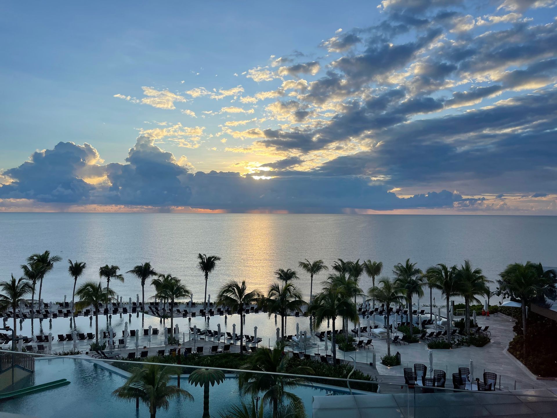 Oceanfront resort at sunset with palm trees and cloudy sky.