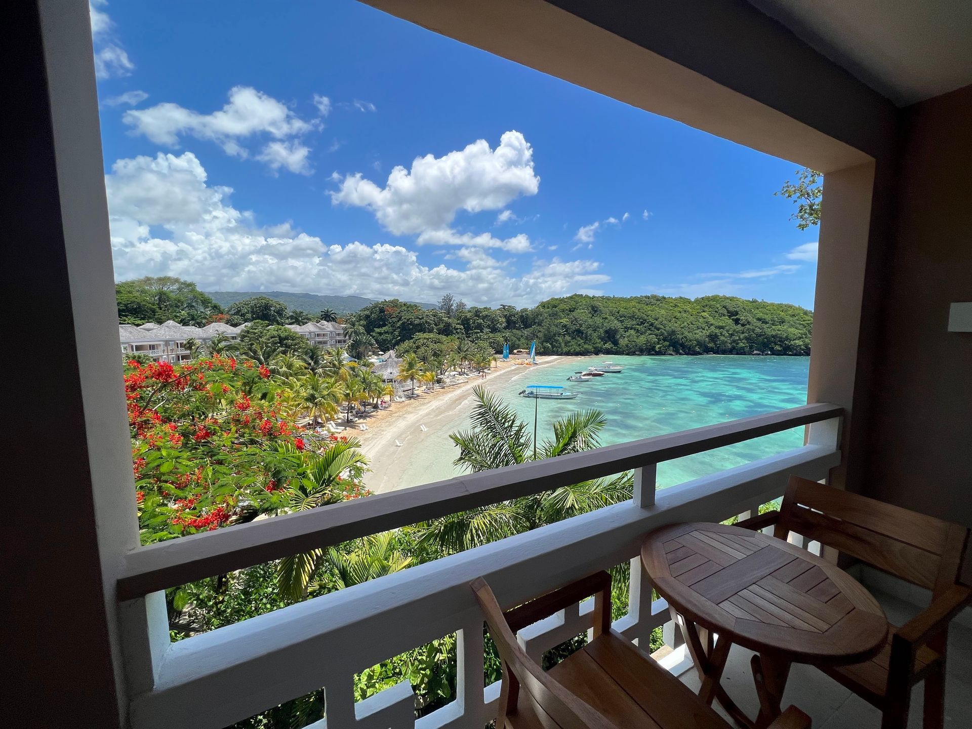 Balcony overlooking turquoise ocean and beach. Wooden furniture on the balcony. Lush greenery and blue sky.