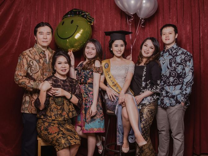 Group posing at a graduation celebration in front of red drapes and balloons, with a large green smiley balloon.