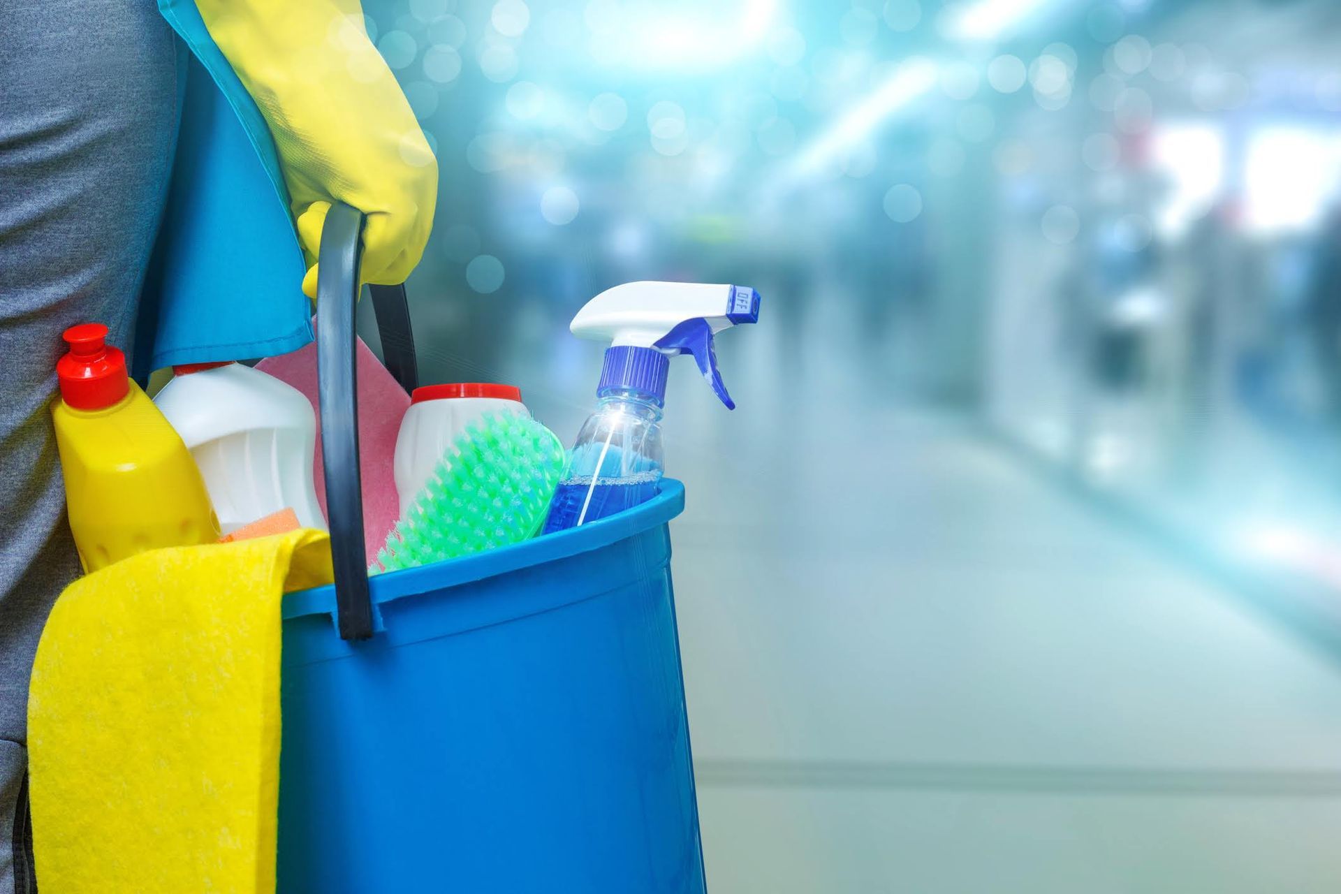 Persona che tiene in mano un secchio blu pieno di prodotti per la pulizia, in un ambiente interno sfocato.