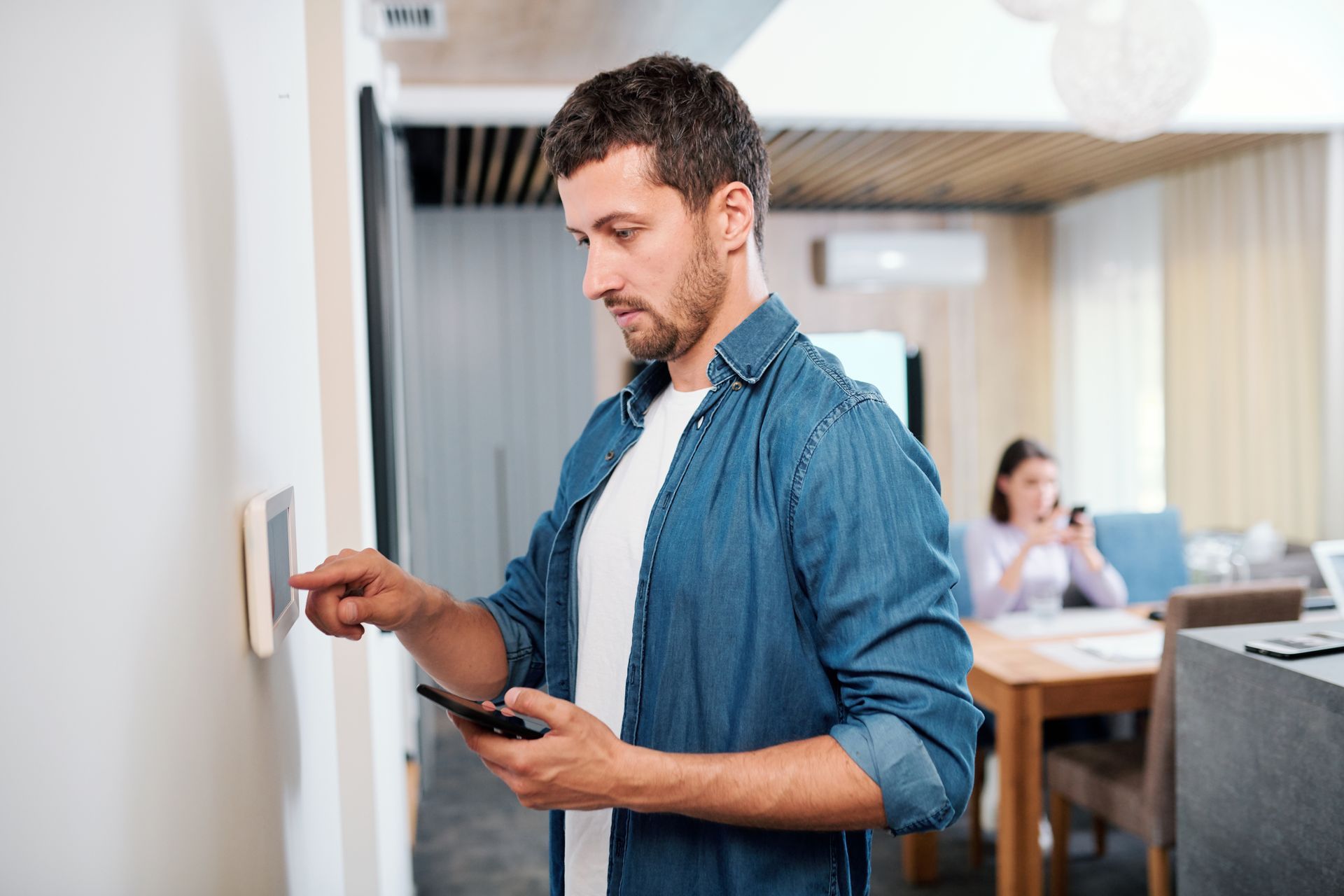 Man using a smart thermostat on a wall, holding a phone. A person is seated at a table in the background.