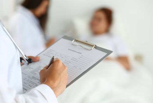 Doctor filling out a clipboard form while a patient lies blurred in a hospital bed