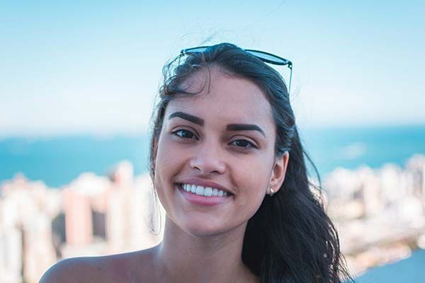 Smiling woman with sunglasses on her head, with a blurred city skyline and ocean behind her.