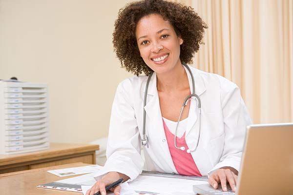 Smiling doctor in a white coat at a desk with a stethoscope and laptop in a bright office