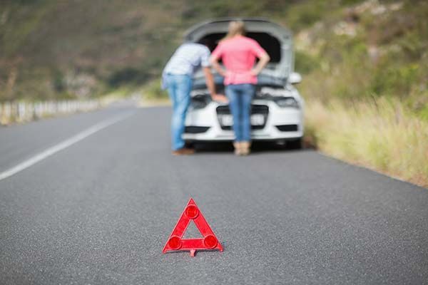 Red warning triangle on road; blurred car with open hood and two people inspecting it in the background