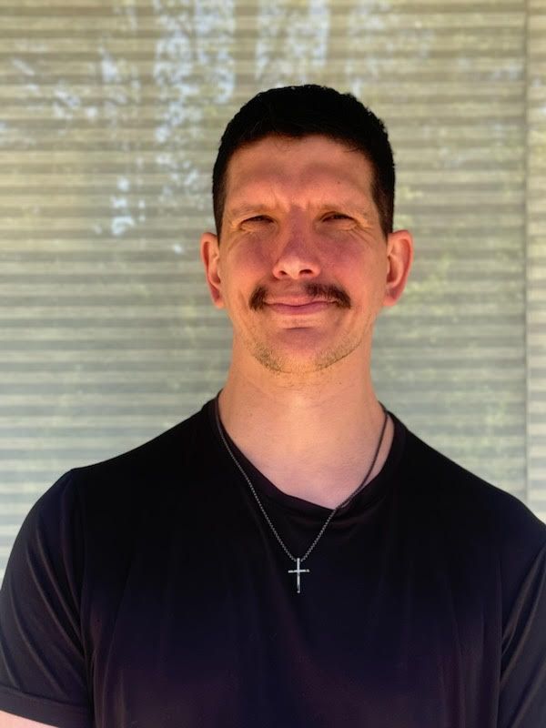 A man with a mustache is wearing a black shirt and a cross necklace.