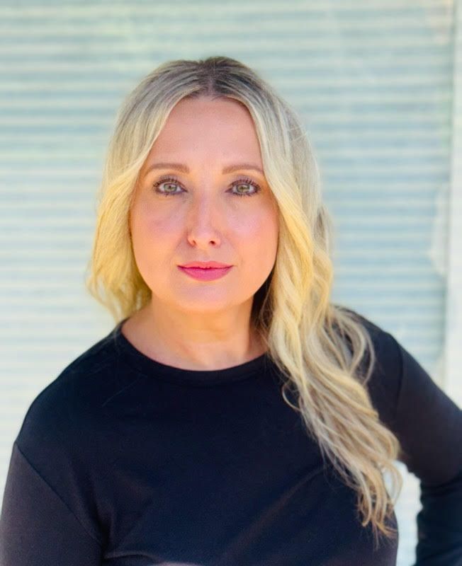 A woman with blonde hair and a black shirt is standing in front of a window.