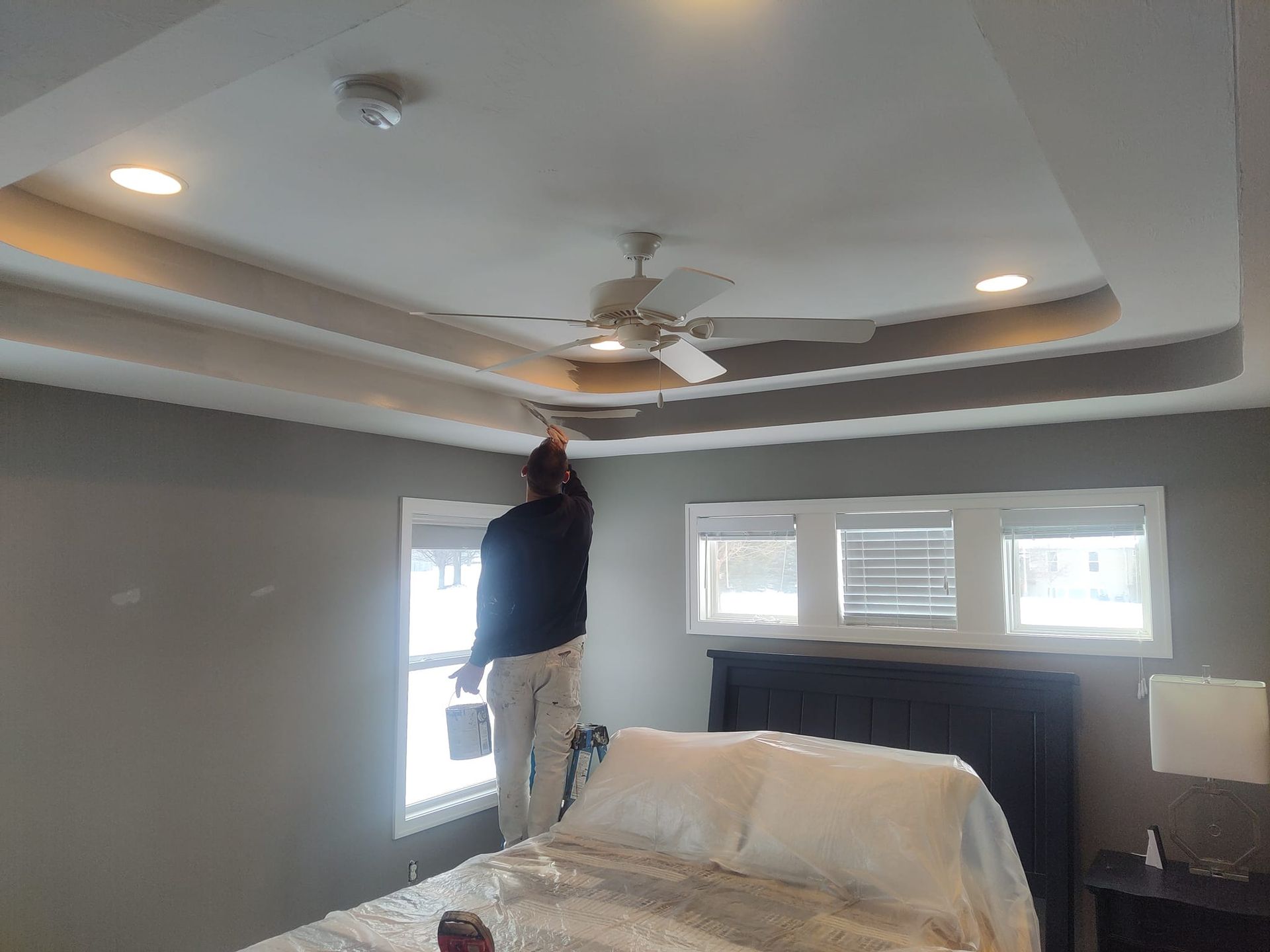 A man is painting the ceiling of a bedroom with a ceiling fan.