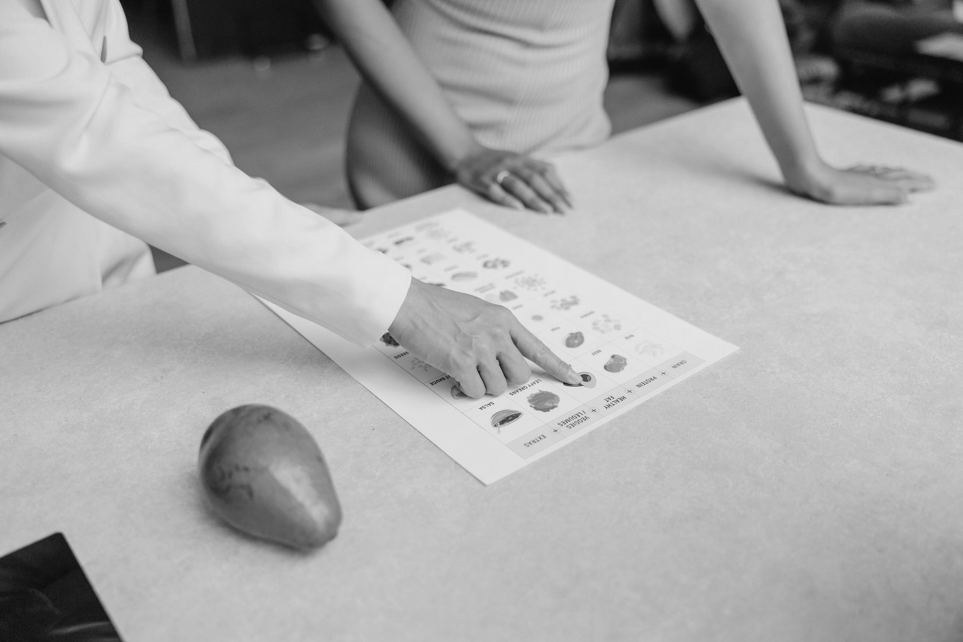 A woman is pointing at a piece of paper on a table next to an avocado.