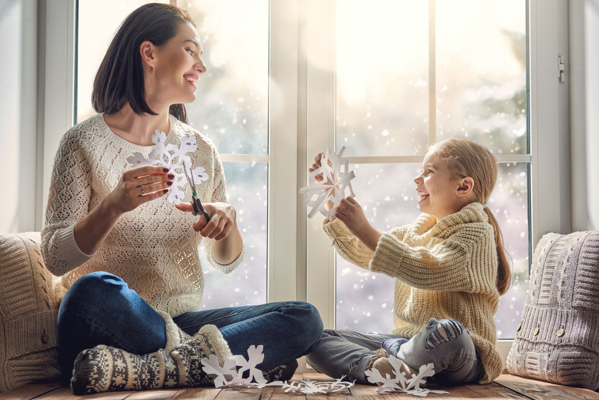 a woman and a little girl are sitting on a window sill making snowflakes .