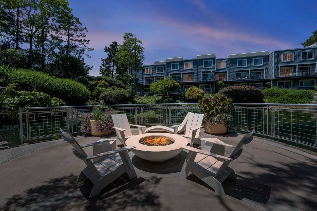 Outdoor communal fire pit surrounded by chairs on a deck, with apartment buildings in the background.
