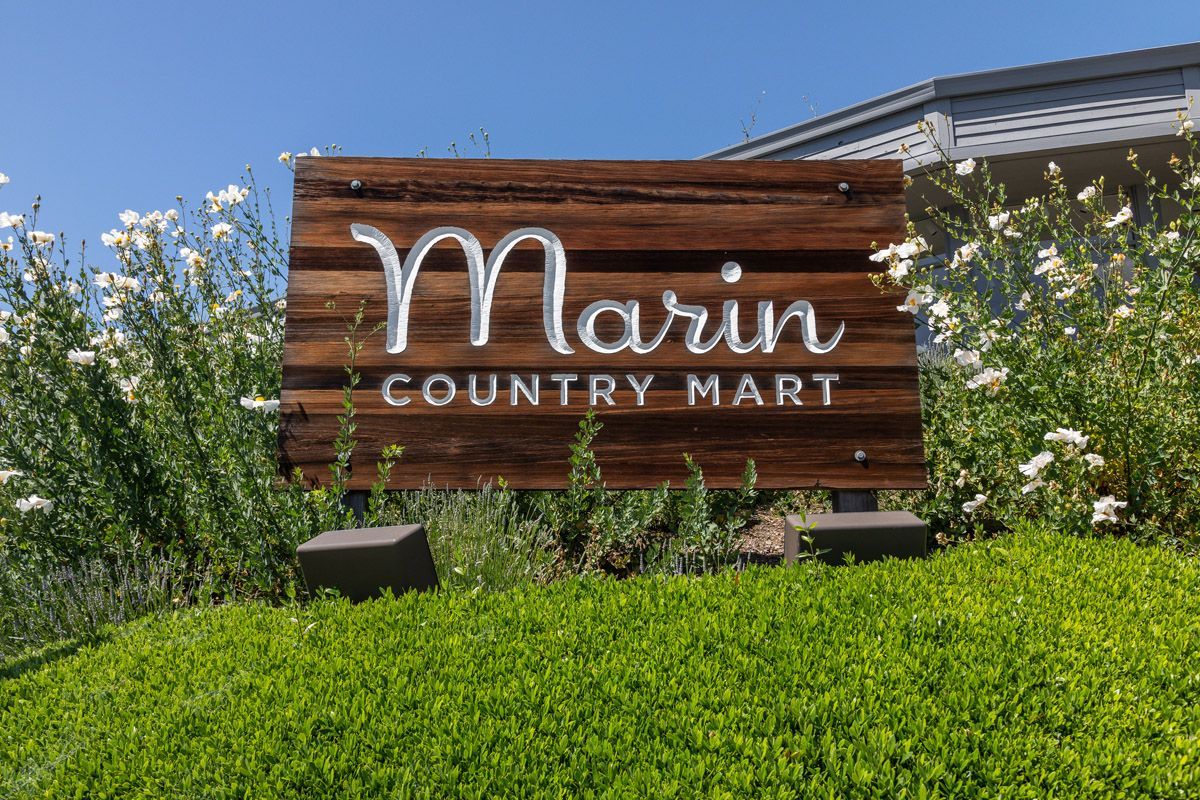 Wooden sign reading 'Marin Country Mart' with shrubs and white flowers against a blue sky.