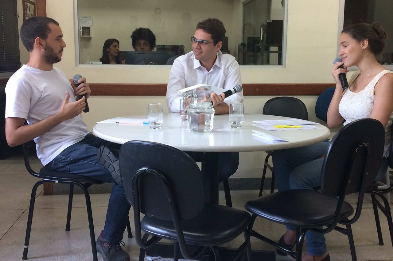 Aula de jornalismo na rádio da faculdade. Danilo se senta à mesa com o jornalista Paulo Henrique Azeredo e sua colega Raissa. Os três estão de blusa branca e microfones nas mãos. Há uma janela de vidro atrás dos três onde é possível ver uma funcionária e uma estagiária cuidando da parte técnica do áudio.