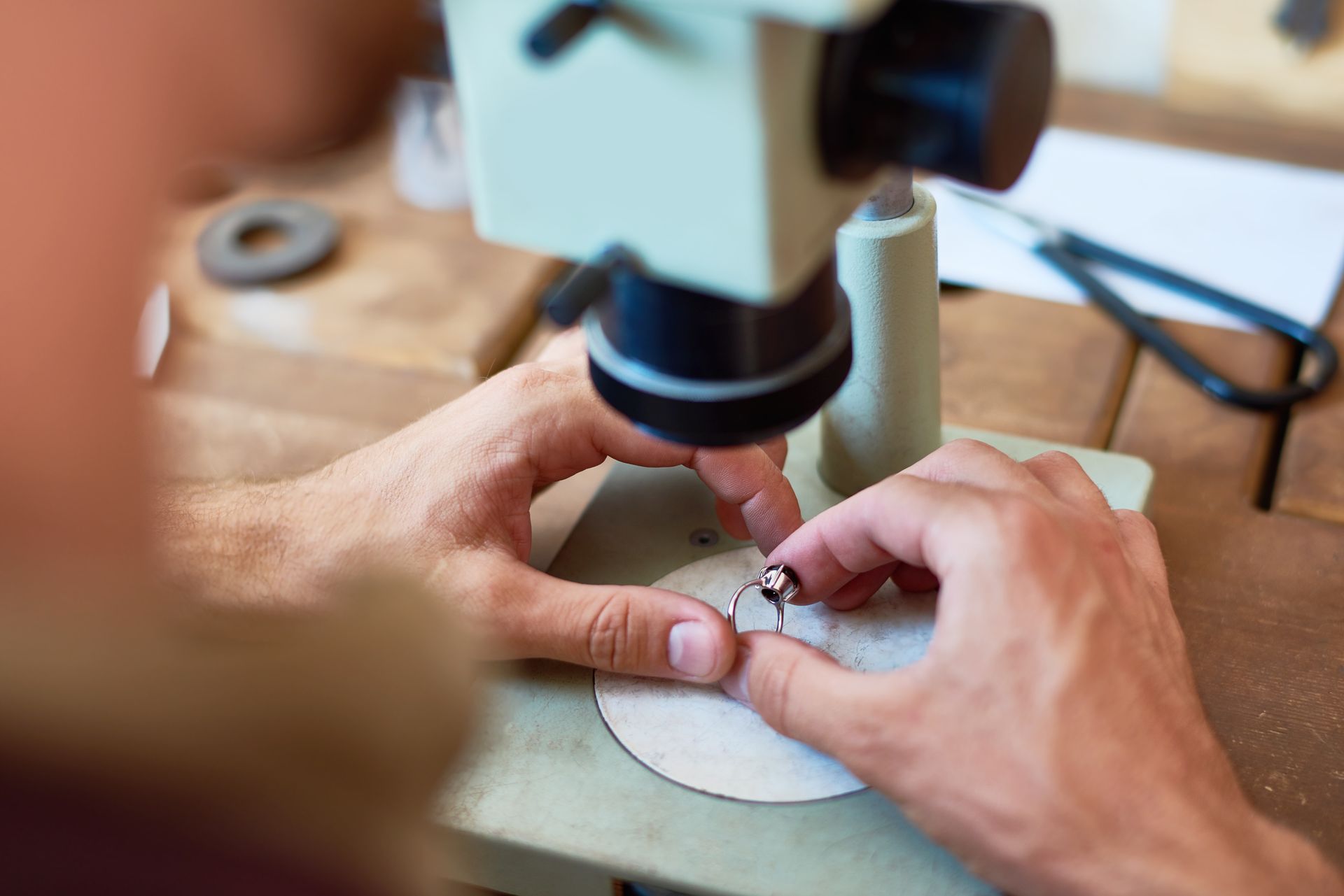 Des mains travaillant sur une bague sous un microscope, sur une surface en bois.