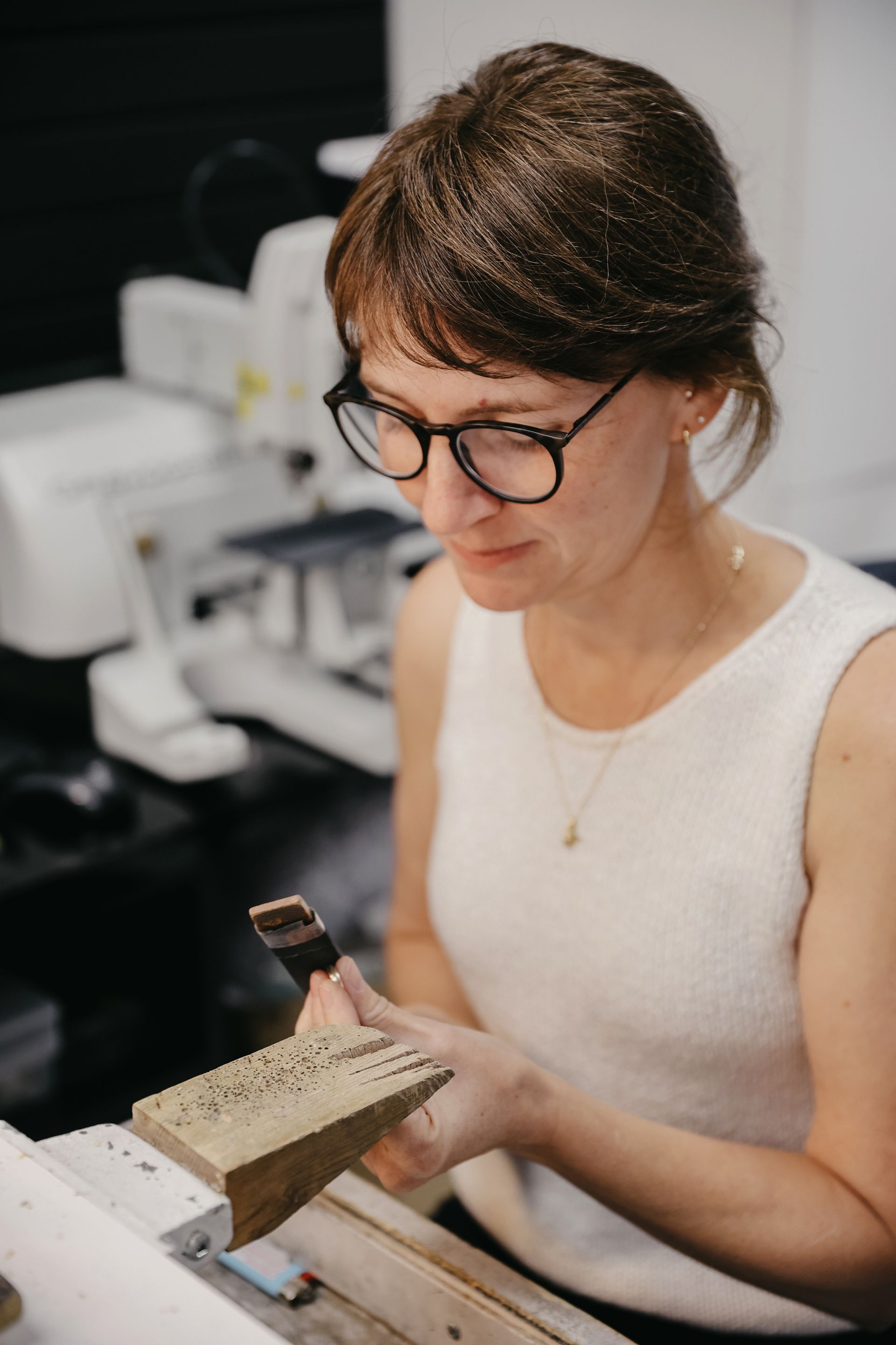 Une femme portant des lunettes examine à la loupe une pièce de métal perforée, près d'une machine.
