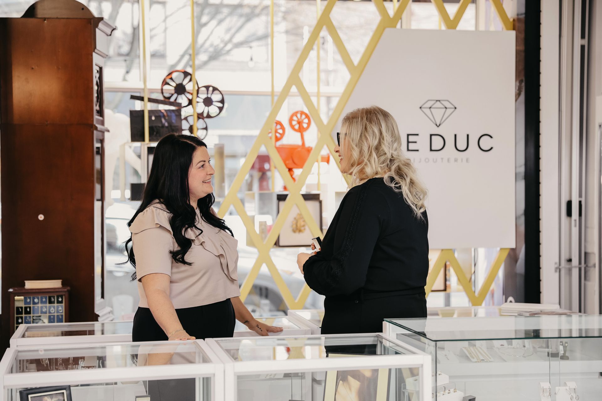 Deux femmes discutent dans une bijouterie, l'une derrière le comptoir. Enseigne ornée d'un logo en forme de diamant.