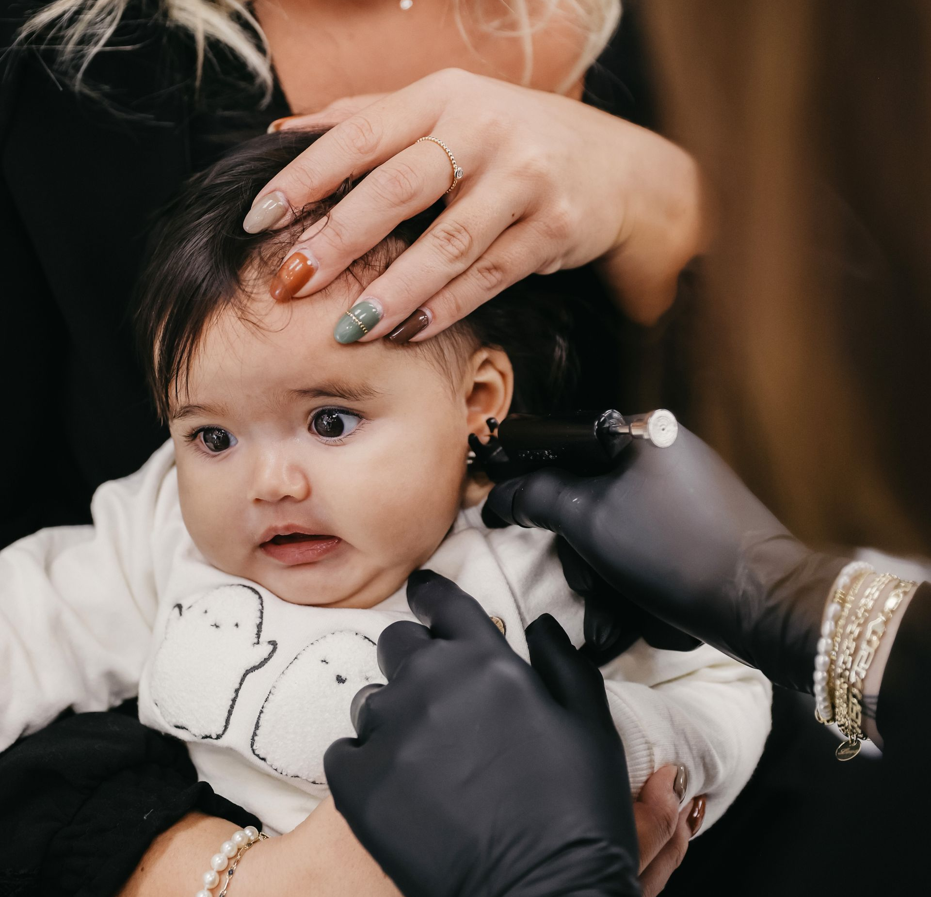 Un bébé se fait percer l'oreille, tenu par quelqu'un, une autre personne effectuant le perçage avec un outil.