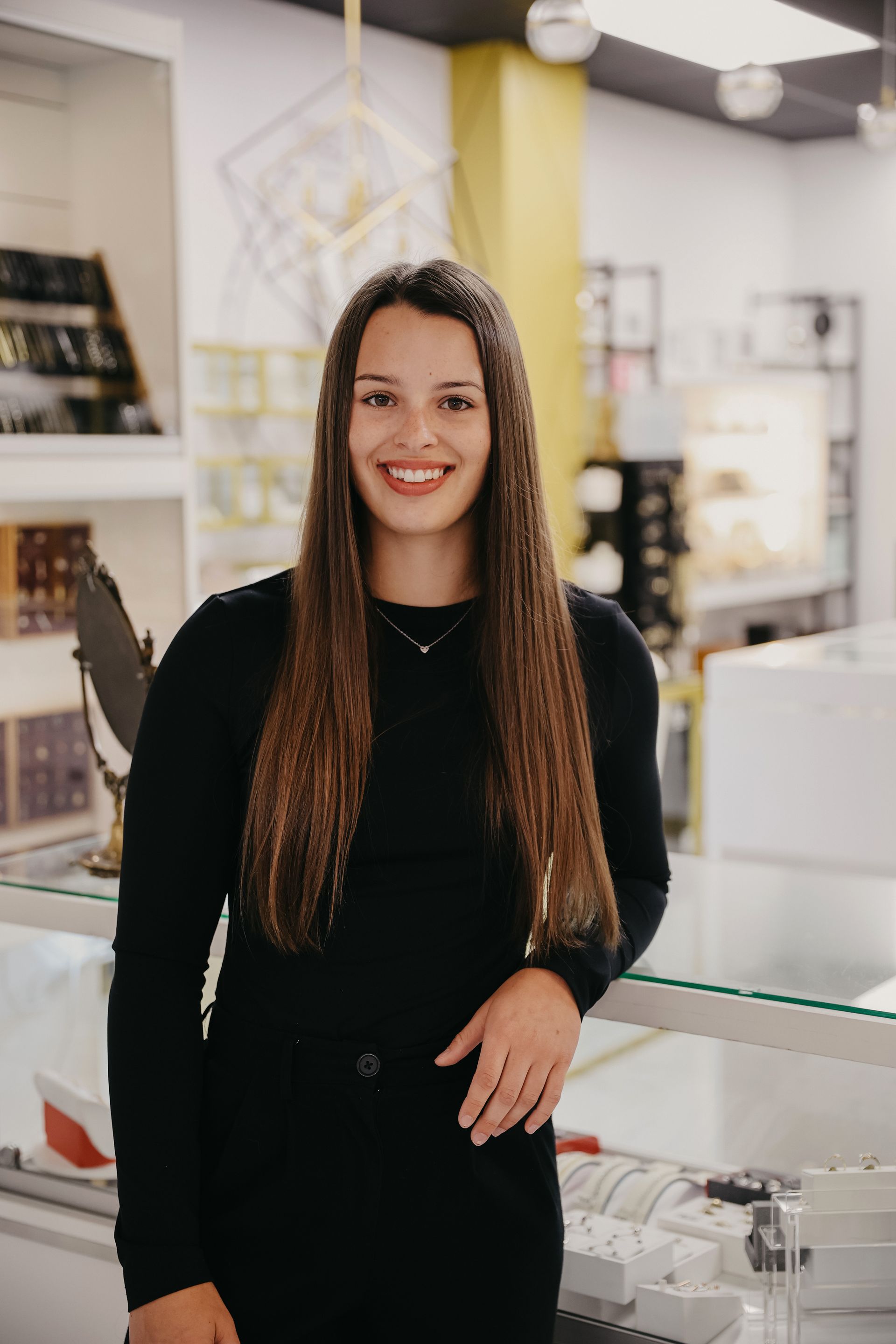 Femme aux longs cheveux bruns souriant dans une bijouterie, vêtue d'un haut noir.