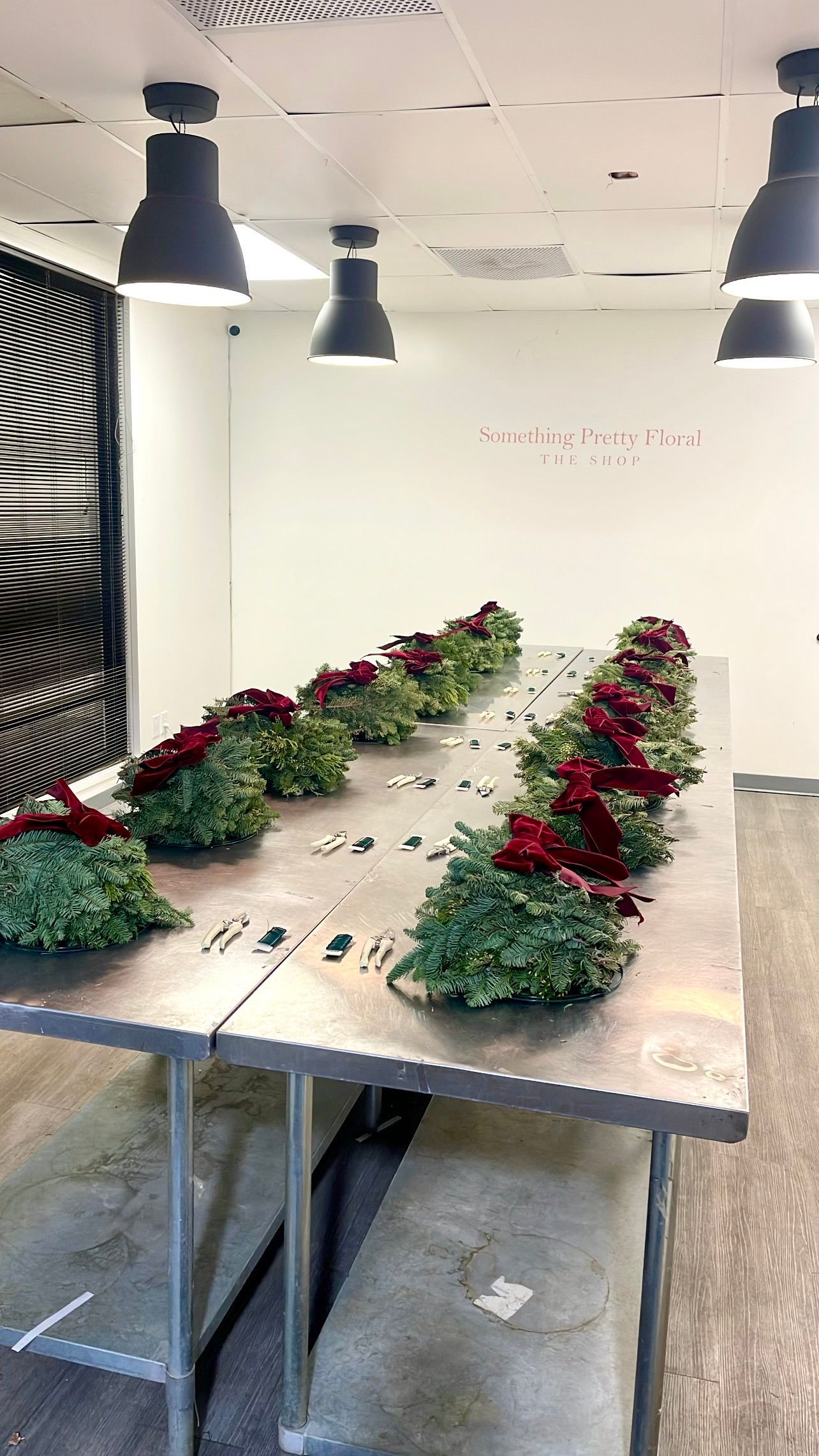 Rows of floral arrangements with red roses on a long metal table beneath pendant lights.