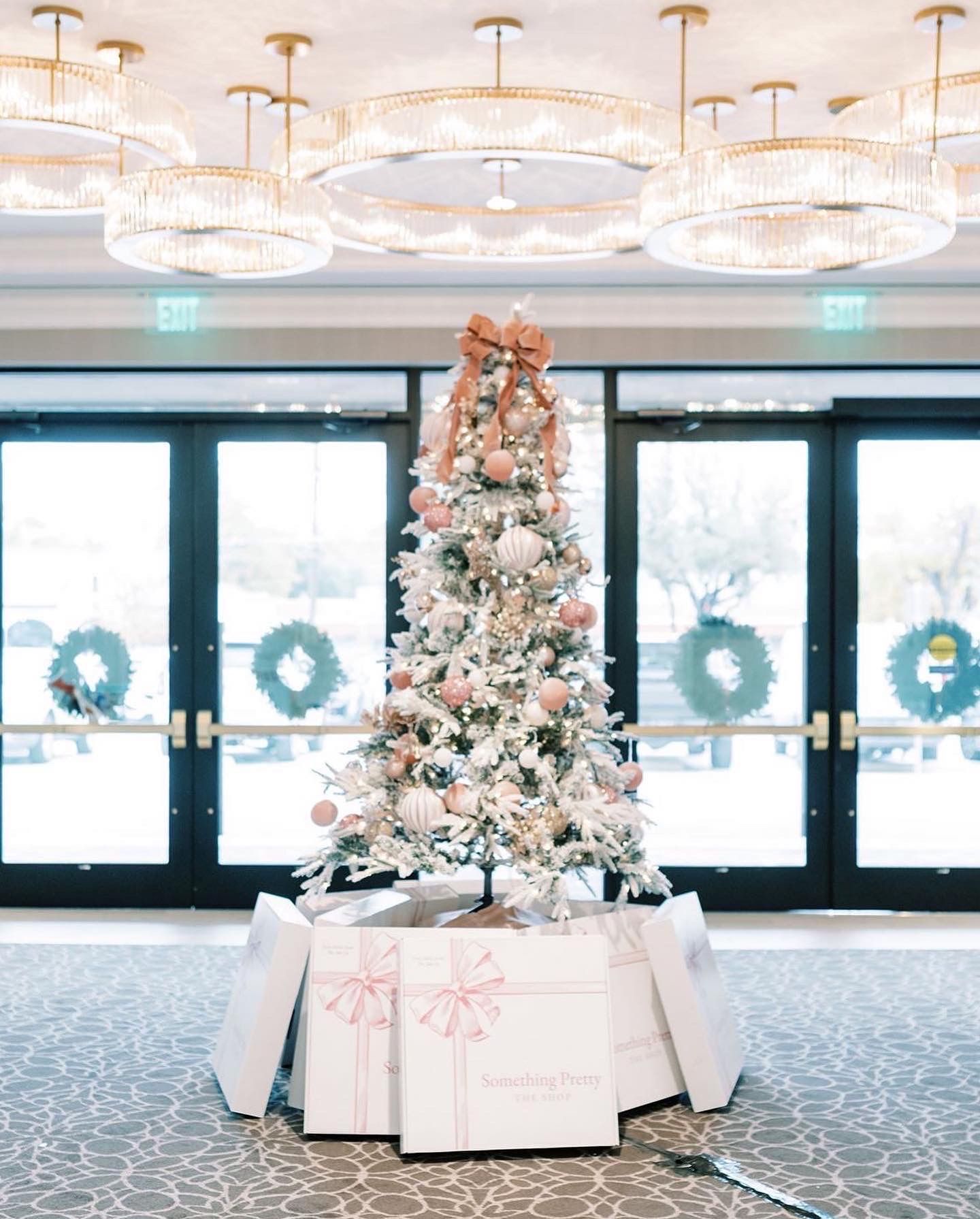 Christmas tree with pink ornaments and bow, surrounded by gift boxes, in a bright lobby.