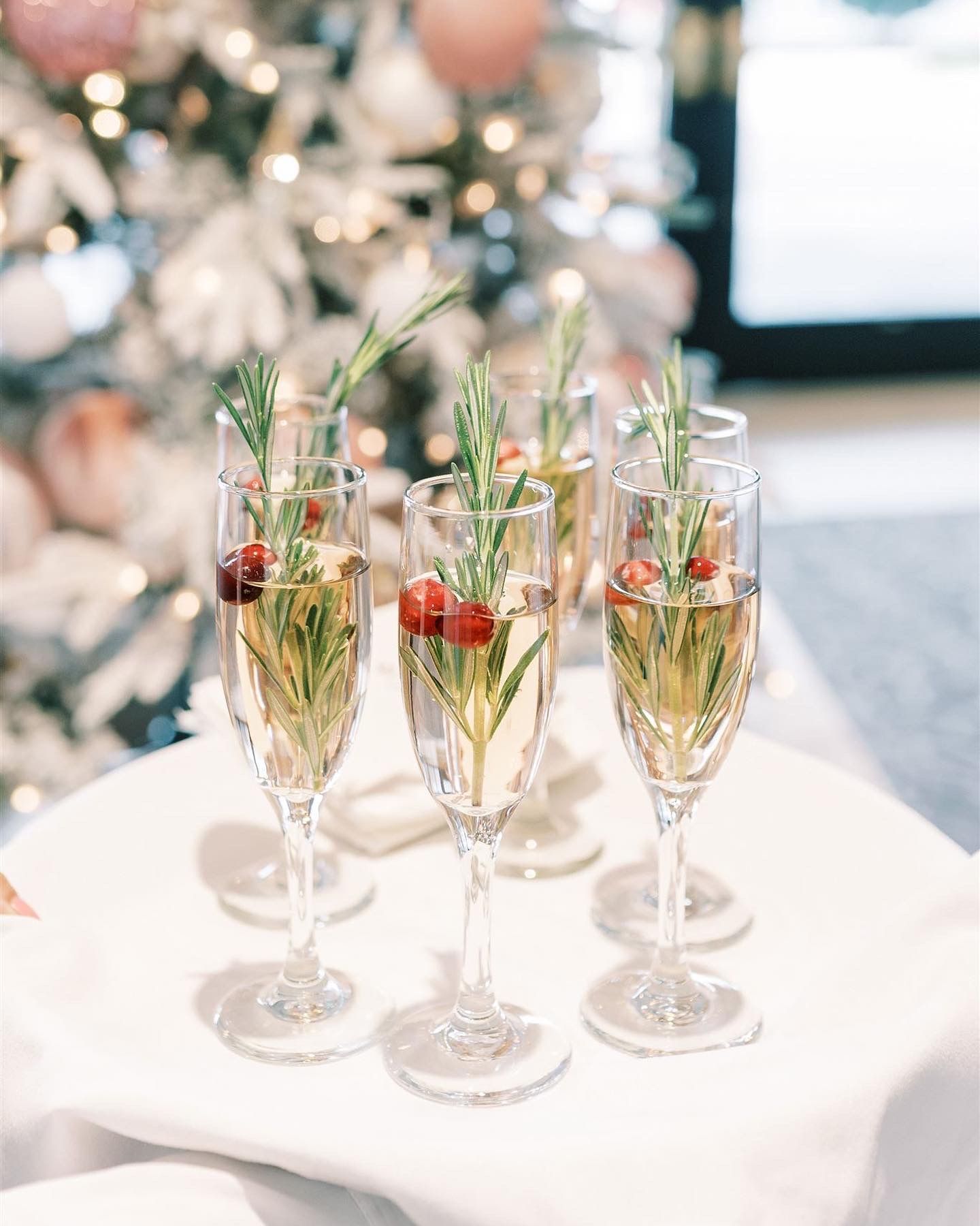 Champagne flutes with cranberries and rosemary sprigs on a white tray, in front of a decorated Christmas tree.