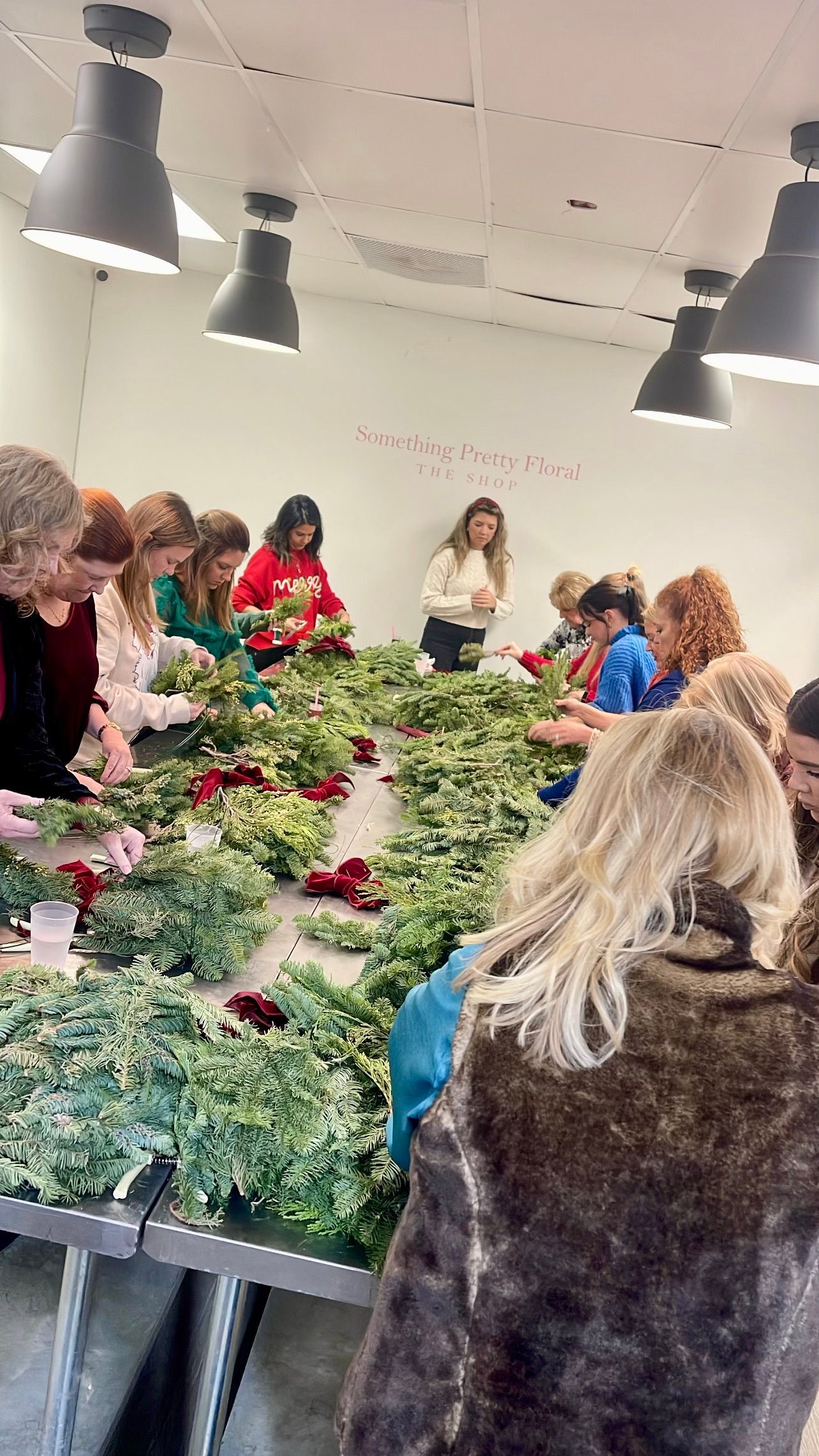 People making wreaths at a long table, indoors with overhead lighting.