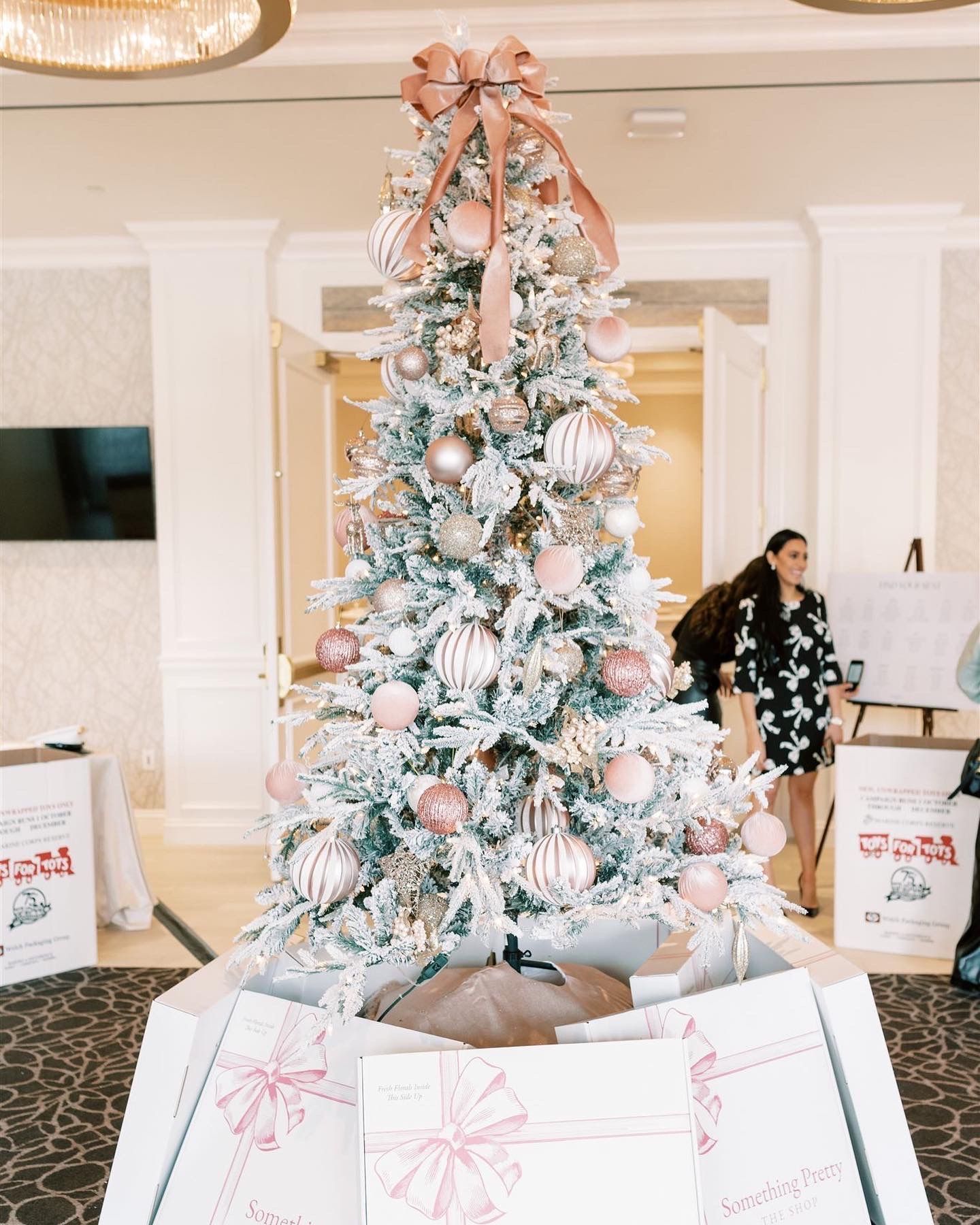 Decorated Christmas tree with pink ornaments and bow, surrounded by gifts. Woman in floral dress in background.