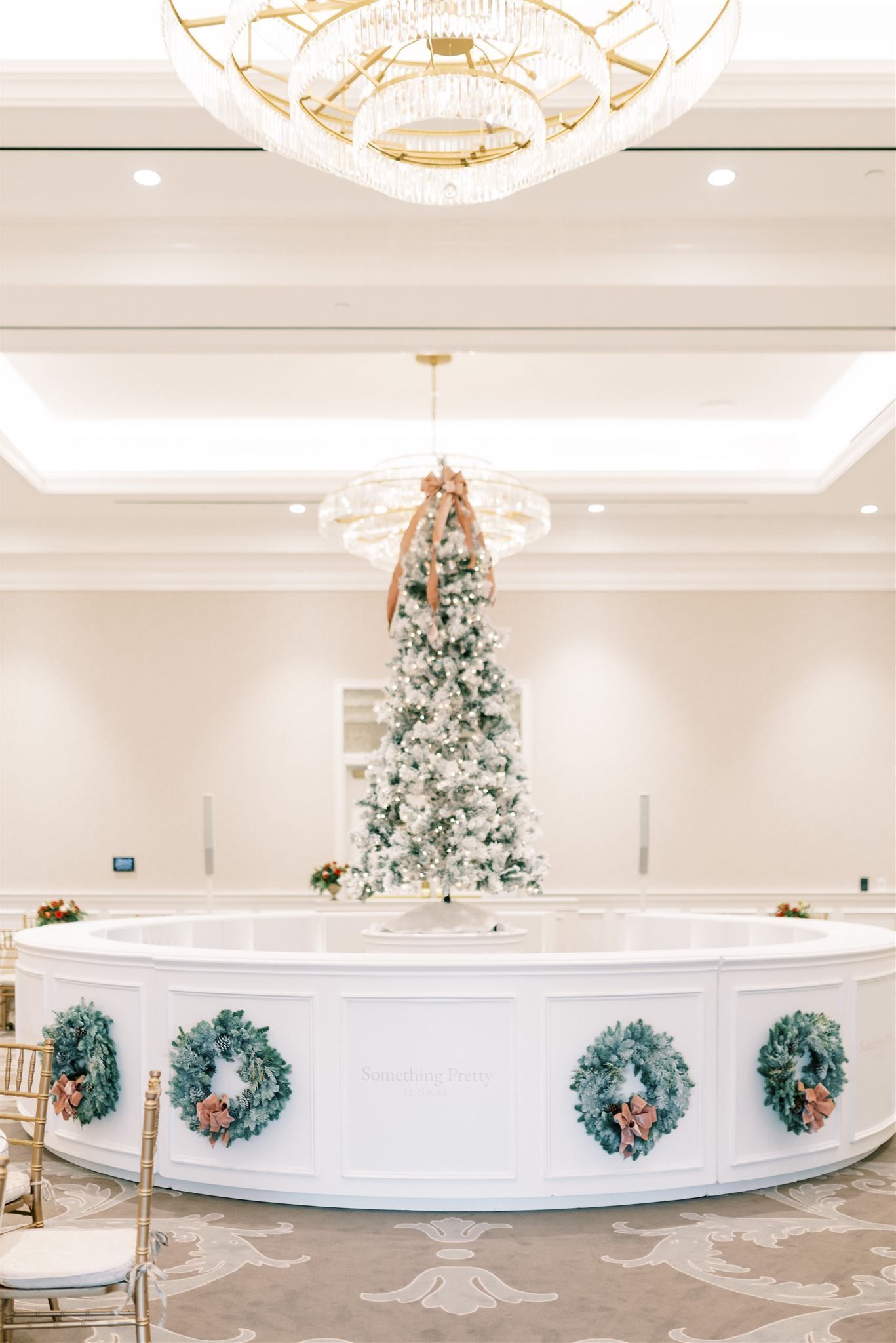 White baptismal font decorated with wreaths and Christmas tree, ornate chandelier in a light, elegant room.