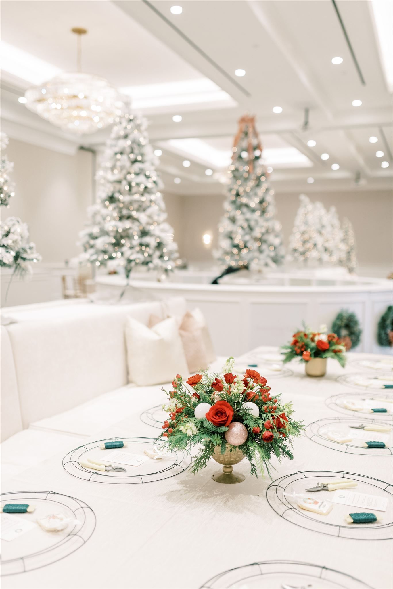 Elegant holiday table setting with floral centerpiece, snowy Christmas trees, and white seating in a bright room.